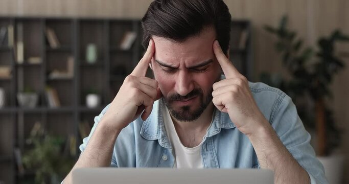 Young man working at office desk closed his eyes rub temples to reduce pain, suffering from chronic severe headache, due long usage of laptop. Troubles at work, thinking process, search ideas concept