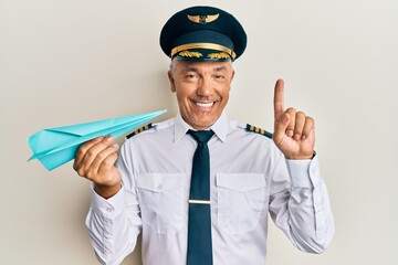 Handsome middle age mature man wearing airplane pilot uniform holding paper plane smiling with an...