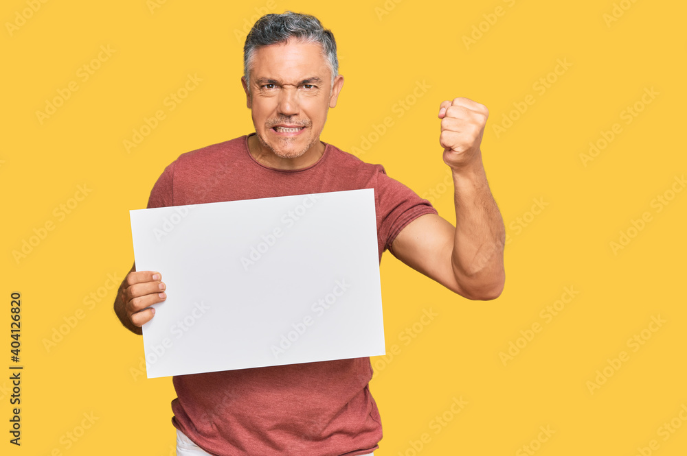 Wall mural handsome middle age man holding blank empty banner annoyed and frustrated shouting with anger, yelli