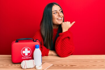 Beautiful hispanic woman with first aid kit pointing thumb up to the side smiling happy with open...
