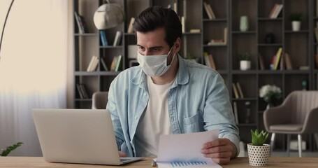 Businessman in surgical protective facemask analyzing financial stats infographic information sit at workplace. Man holds document with charts and graphs, prepare report, make forecast or presentation - Powered by Adobe