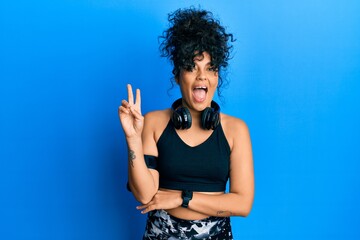 Young hispanic woman wearing gym clothes and using headphones smiling with happy face winking at...