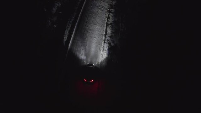 Car or vehicle driving on a snowy road in the winter at night. Headlights lighting up the icy road in the dark. Heading away to distance. Following or tracking aerial drone shot.