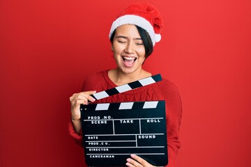 Beautiful young woman with short hair wearing christmas hat holding clapboard smiling and laughing...