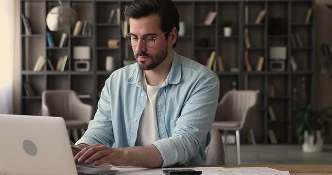 Focused serious man sit at desk working calculate costs using calculator laptop app, pay through e-bank application. Business, household expenses, accounting, managing finances, check incomes concept