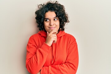 Fototapeta na wymiar Young hispanic woman with curly hair wearing casual sweatshirt with hand on chin thinking about question, pensive expression. smiling with thoughtful face. doubt concept.