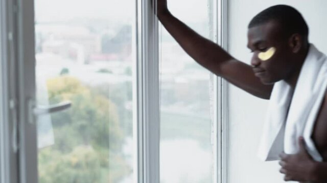 New day. Fresh start. Inspiration contemplation. Confident curious happy shirtless African man with gold hydrogel eye patches white bath towel smiling looking through window on light background.
