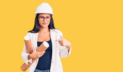 Young beautiful latin girl wearing architect hardhat holding blueprints smiling happy pointing with...