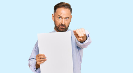 Handsome middle age man holding blank empty banner pointing with finger to the camera and to you, confident gesture looking serious