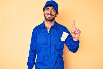 Handsome young man with curly hair and bear wearing builder jumpsuit uniform pointing finger up with successful idea. exited and happy. number one.