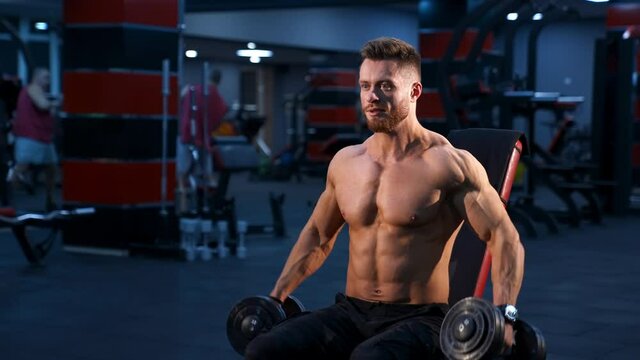 Handsome power athletic man bodybuilder doing exercises with dumbbells. Fitness muscular body on dark gym background.
