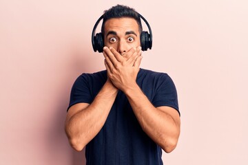 Young hispanic man listening to music using headphones shocked covering mouth with hands for...