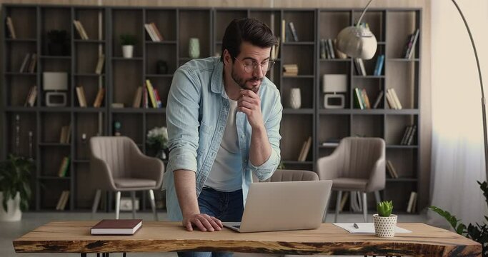 Thoughtful thinking businessman standing in office lean over desk hurrying make urgent task working on laptop. Company employee planning, considering research results, business project using computer