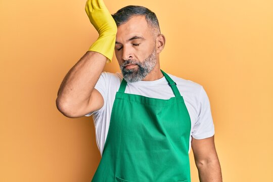 Middle age handsome man wearing cleaner apron and gloves surprised with hand on head for mistake, remember error. forgot, bad memory concept.