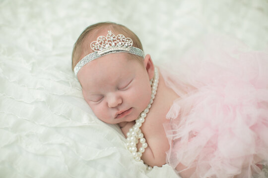 Close up of a sweet newborn infant baby girl laying on a cream-colored neutral background with a crown on her head like a little princess royalty.