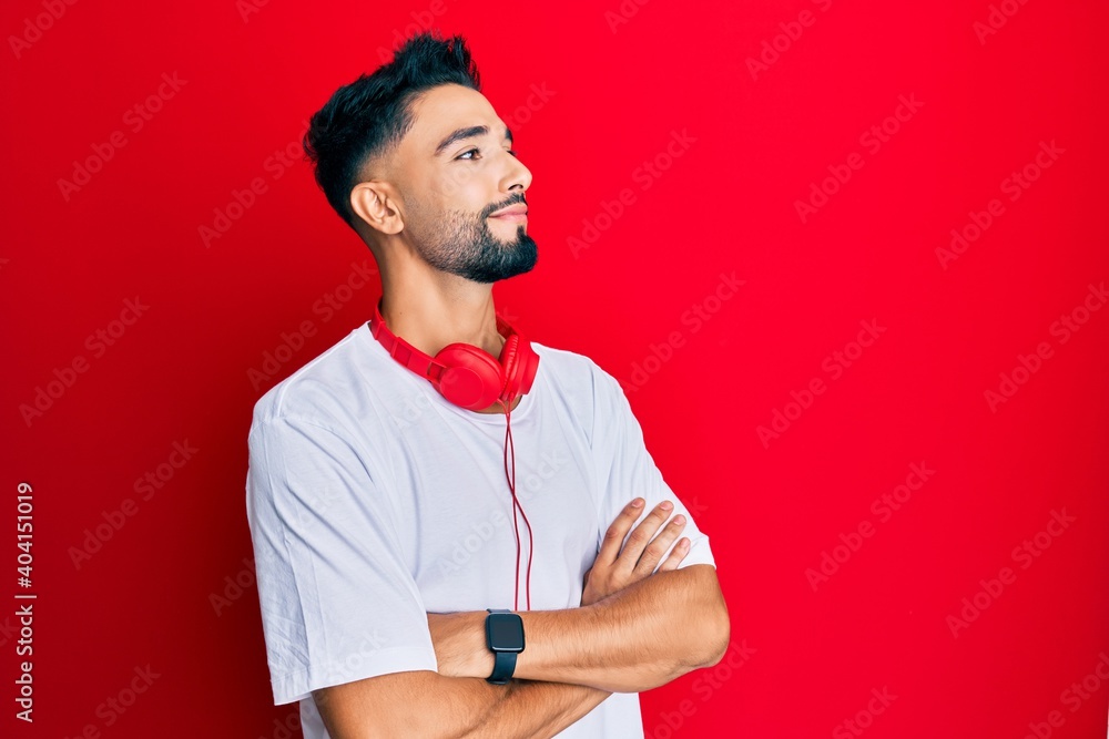 Wall mural Young man with beard listening to music using headphones looking to the side with arms crossed convinced and confident