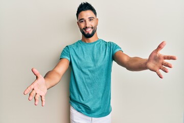 Young man with beard wearing casual clothes looking at the camera smiling with open arms for hug. cheerful expression embracing happiness. - obrazy, fototapety, plakaty