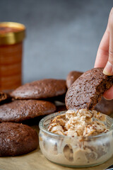 Woman's hand picking up a fluffy chocolate brownie cookie dipped in a glass bowl of peanut butter ice cream. Healthy homemade gluten free chocolate chip cookies in the background with ice cream bucket