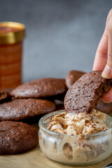 Woman's hand picking up a fluffy chocolate brownie cookie dipped in a glass bowl of peanut butter ice cream. Healthy homemade gluten free chocolate chip cookies in the background with ice cream bucket