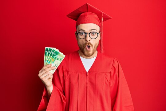 Young redhead man wearing graduation robe holding 50 shekels scared and amazed with open mouth for surprise, disbelief face