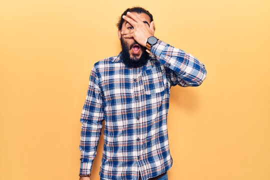 Young arab man wearing casual clothes peeking in shock covering face and eyes with hand, looking through fingers afraid