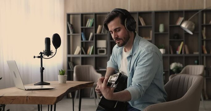 Serious focused man guitarist wear wireless headphones playing a guitar seated in domestic cozy room in front of opened laptop, lead on-line class, teach learner, using online lesson. Hobby concept