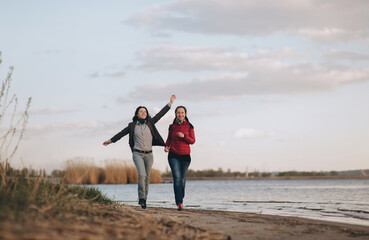 Two girlfriends in blue jeans, a jacket and a shirt run along the river bank in the evening of sunset and laugh with joy. The concept of a spring walk outdoors.