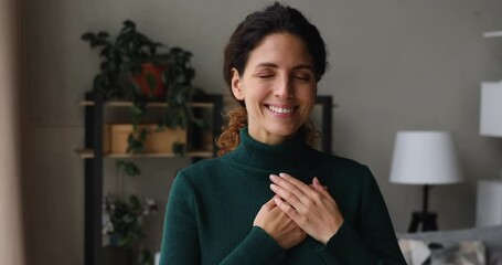 Smiling benevolent beautiful sincere woman standing indoor with eyes closed put folded palms on heart feeling deep gratitude and thankful. Express appreciation sign, hope, believe and charity concept