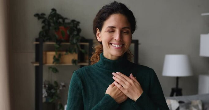 Smiling benevolent beautiful sincere woman standing indoor with eyes closed put folded palms on heart feeling deep gratitude and thankful. Express appreciation sign, hope, believe and charity concept