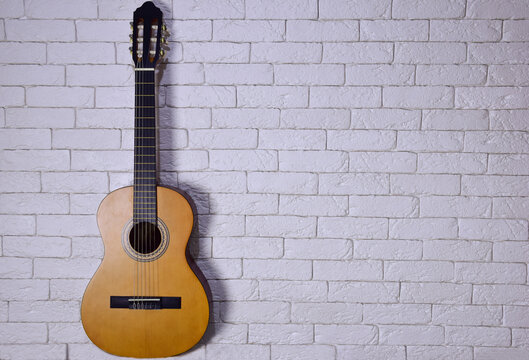 An acoustic guitar stands upright against a light brick wall on the left side. Plenty of space on the right for caption or text © Serhii