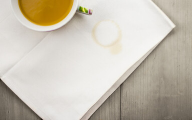 White cotton napkin with a round light brown coffee stain, on a wooden grey background, ceramic vintage cup with coffee and milk, top view