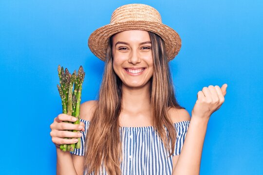 Young beautiful girl wearing hat holding asparagus screaming proud, celebrating victory and success very excited with raised arm