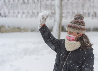 Side view of a little girl wearing a coronavirus face mask throwing a snowball during a snowfall