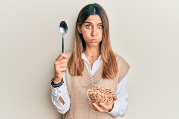 Brunette young woman eating healthy whole grain cereals with spoon puffing cheeks with funny face....