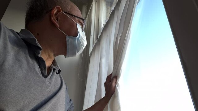 A Chinese elderly man self isolating at home wearing a face mask and looking out of the window of a sunny day with longing for the outdoors.