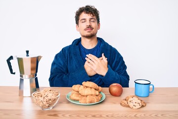 Young caucasian man with curly hair sitting on the table having breakfast smiling with hands on chest, eyes closed with grateful gesture on face. health concept.