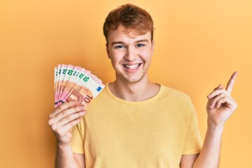 Young caucasian man holding bunch of 10 euro banknotes smiling happy pointing with hand and finger to the side