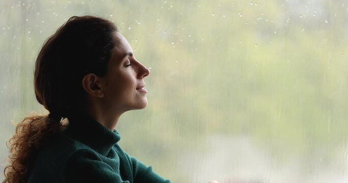 Close up profile face calm woman sit on windowsill near window closed eyes enjoy fresh air, do breathing exercise, relaxing indoor, rainy weather outside. No stress, meditation, anxiety relief concept
