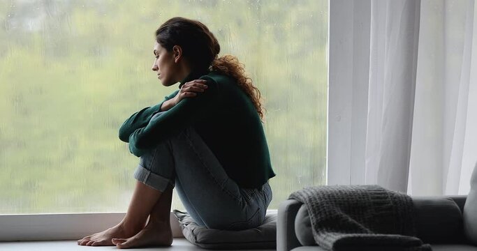 Sad woman sit barefoot on window sill looks out window at rainy gloomy weather day feels lonely and melancholic, thinks about personal life troubles, break up divorce. Solitude, autumn blues concept