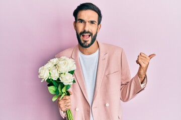 Young hispanic man holding flowers pointing thumb up to the side smiling happy with open mouth