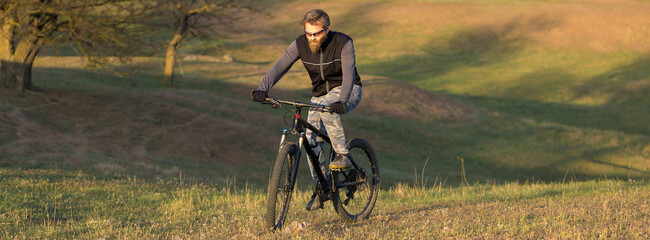 Fototapeta premium Sports brutal bearded guy on a modern mountain bike. Cyclist on the green hills in the spring.