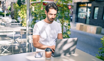 Young hispanic man with serious expression working using laptop at coffee shop terrace.