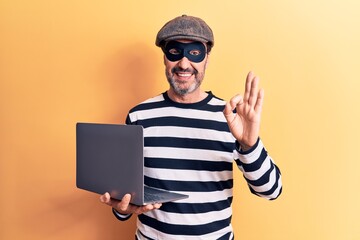 Middle age handsome burglar man wearing cap and mask using laptop over yellow background doing ok...
