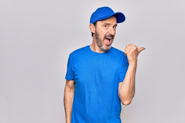 Middle age handsome deliveryman wearing cap standing over isolated white background pointing thumb up to the side smiling happy with open mouth