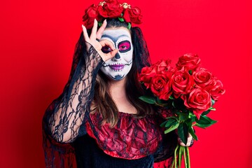 Young woman wearing day of the dead costume holding flowers smiling happy doing ok sign with hand on eye looking through fingers