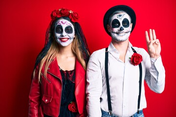 Couple wearing day of the dead costume over red showing and pointing up with fingers number three while smiling confident and happy.