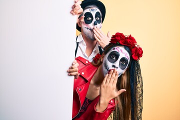 Couple wearing day of the dead costume holding blank empty banner covering mouth with hand, shocked...