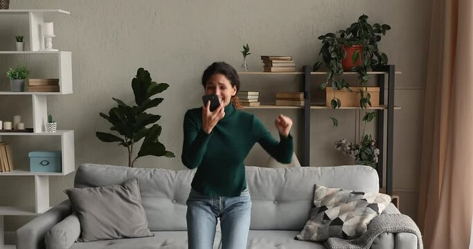 Young woman sit on couch in living room use smartphone read sms, stand up jumps, laughs, dance feels excited looks crazy, very happy. Gambling winner enjoy moment of victory, candid emotions concept