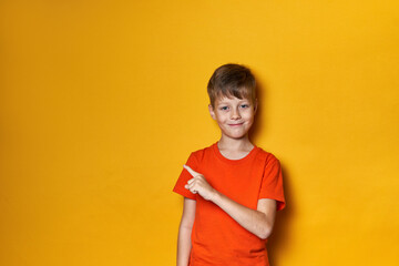 Smiling cute boy in orange t-shirt on a yellow isolated background points his finger to the side at the place for text, banner