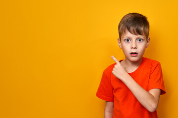 Surprised with open mouth boy in orange t-shirt on yellow isolated background points his finger to...